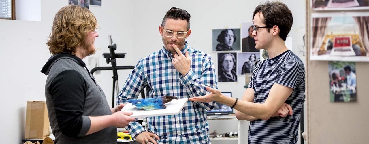 Two male art students consider a piece of art with their professor.